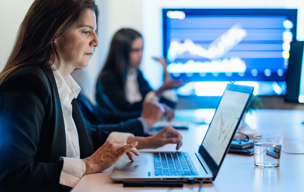 Business professionals in a meeting review data on a laptop while a large monitor shows a stock chart in the background.