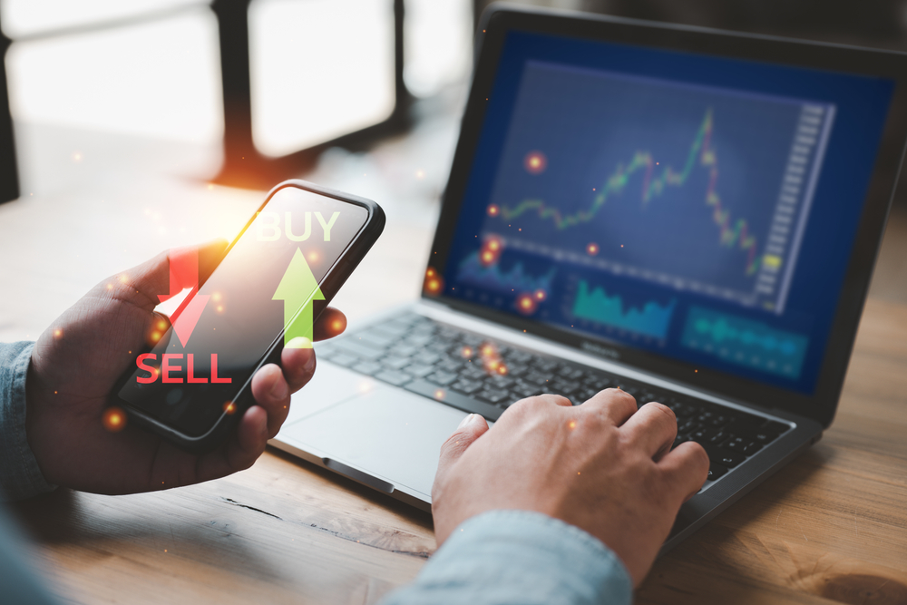 Person holding a smartphone displaying BUY and SELL arrows beside a laptop showing a stock chart.